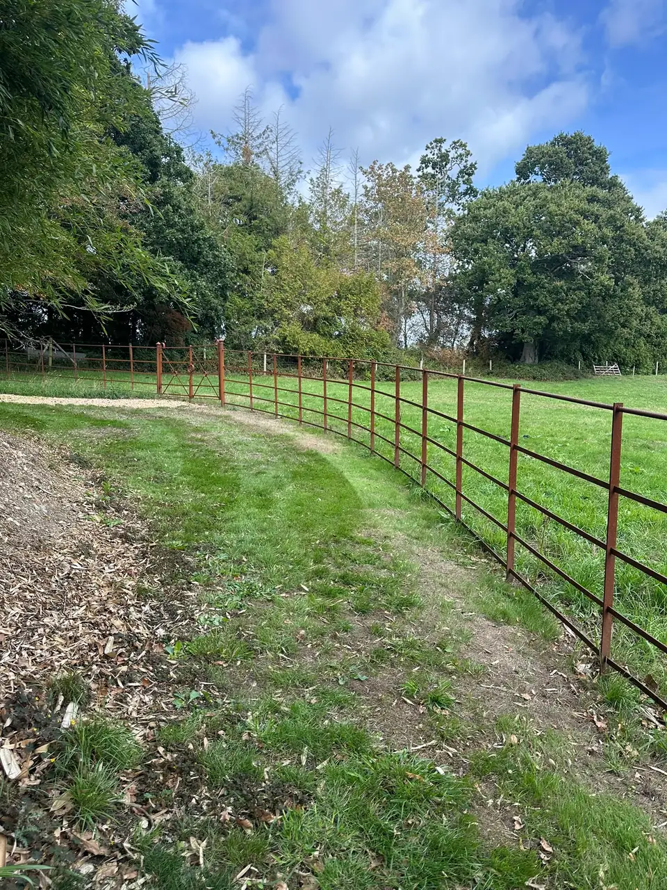 Curved run of oxide-brown estate fencing following a grass track around a paddock, with the gates in the distance